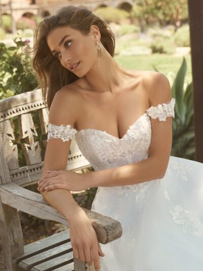 A young woman with long, brown hair sits on a wooden bench, wearing a white, off-the-shoulder wedding dress with a sweetheart neckline. The dress has a lace floral pattern on the bodice and a full skirt. The woman is wearing a ring on her left hand and a pair of gold earrings. She is looking away from the camera with a gentle smile. In the background, there is a stone building and lush greenery.